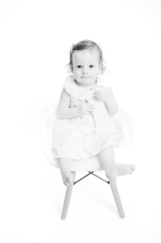 A one-year old girl sitting in a chair, holding a wooden number one and smiling at the camera in black and white